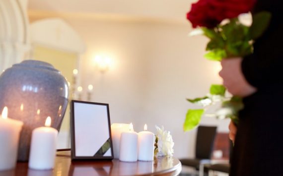 cremation, people and mourning concept -  cinerary urn, photo frame with black ribbon and woman holding red roses at funeral in church